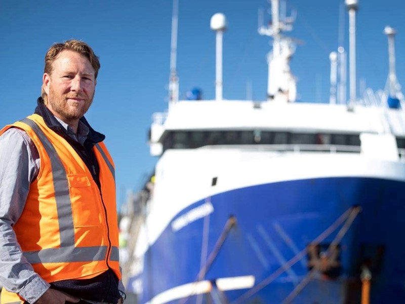 Man wearing high-visibility clothing standing next to a Sealord fishing vessel with a clear blue sky in the background.
