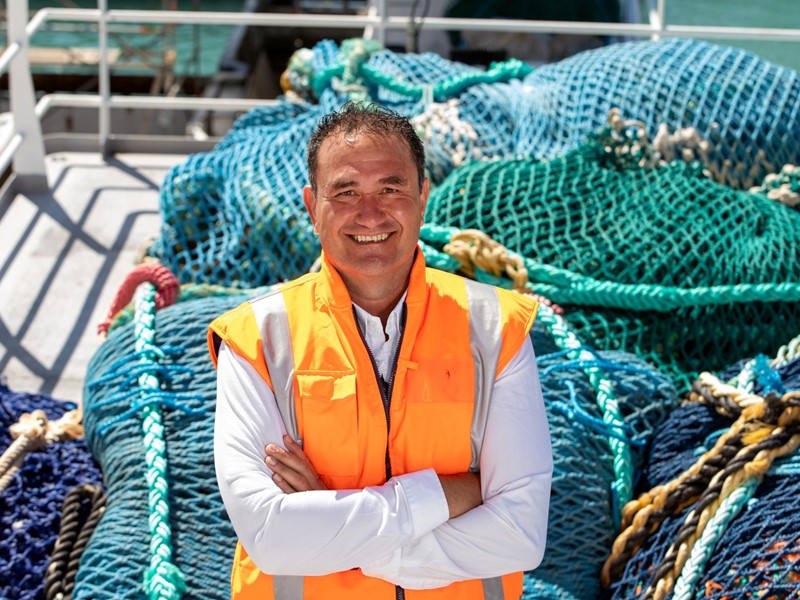 Man in high-visibility gear standing on a boat with a pile of blue and green ropes and fishing equipment behind him.