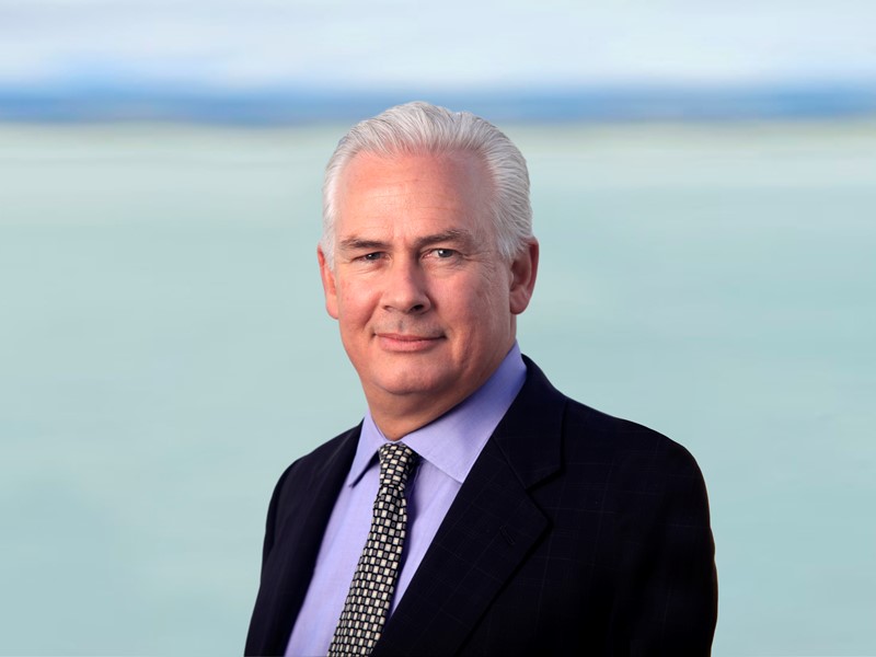 man with gray hair standing infront of the sea and mountains is wearing a purple shirt, patterned tie and black blazer