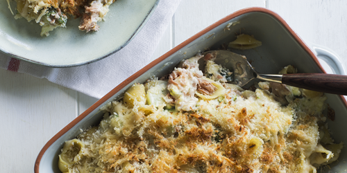 Golden-baked tuna pasta topped with crispy breadcrumbs in a ceramic dish, with creamy tuna, pasta shells, and herbs, served with a portion on a plate nearby.