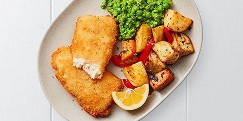 Plate of crispy crumbed hoki fillets with vibrant smashed peas, roasted potatoes and red capsicum, served with a lemon wedge.
