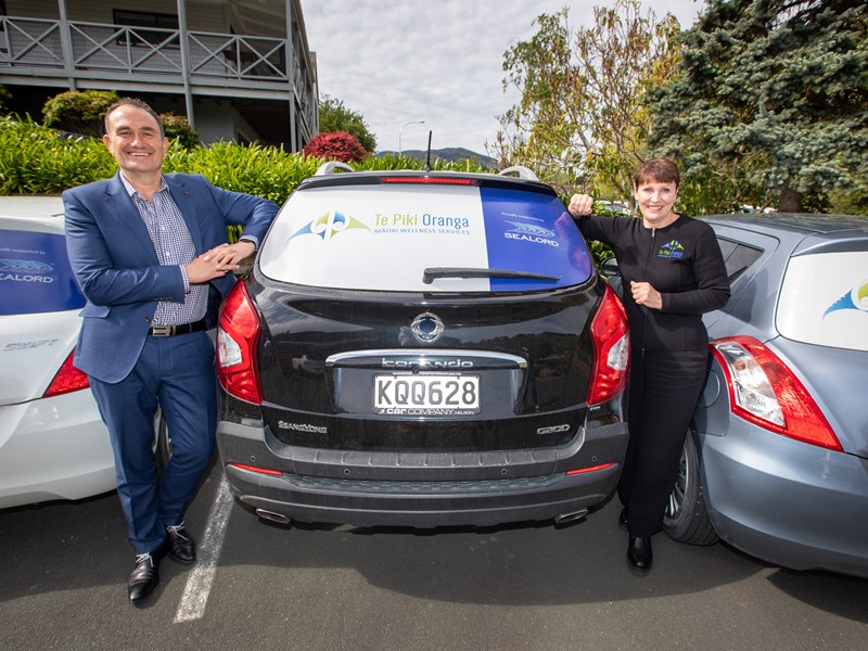 Two people standing in front of the rear window of a parked car displaying the Te Piki Oranga logo, with buildings and a hedge visible in the background. - sealord agreeing to help fund maori health services