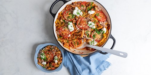 Cheesy baked tuna pasta in a large oven dish, topped with roasted tomato slices, fresh herbs, and dollops of creamy ricotta, served with a side bowl portion.