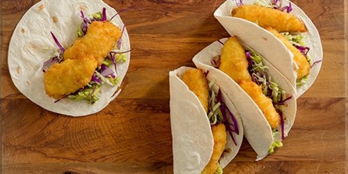 Four soft tacos filled with crumbed fish fillets, shredded cabbage, and slaw on a wooden board.