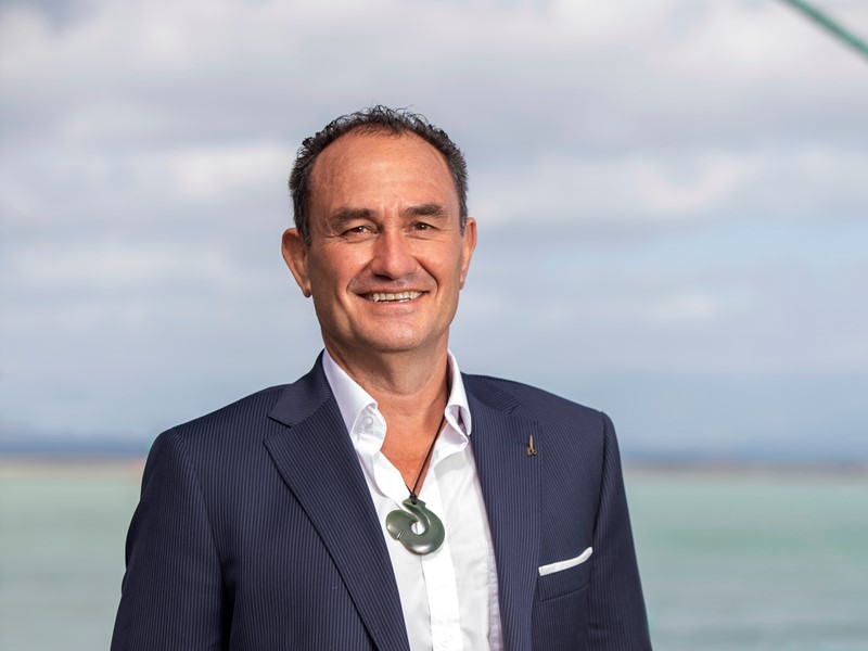 Smiling man in a suit wearing a pounamu (greenstone) pendant, standing outdoors with a coastal landscape in the background.