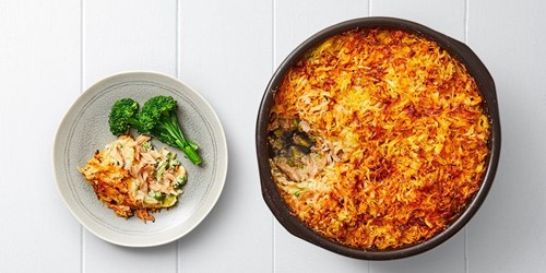 Golden smoked fish pie topped with crispy grated potato and kumara rosti, served with steamed broccolini on a grey plate beside the full pie in a black baking dish.