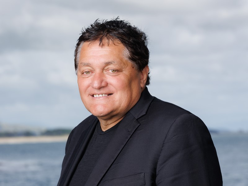 Portrait of a man standing in front of a cloudy sky and sea, with distant land visible in the background.