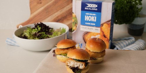 Classic Fish Sliders made with Sealord hoki fillets, placed on a chopping board infront of frozen hoki sealord packaging and next to a white bowl of fresh greens.