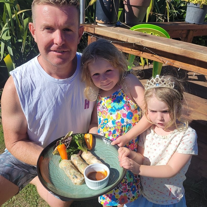 Man and two young girls outdoors holding a plate of homemade rice paper rolls with salad and dipping sauce, enjoying a sunny day together.