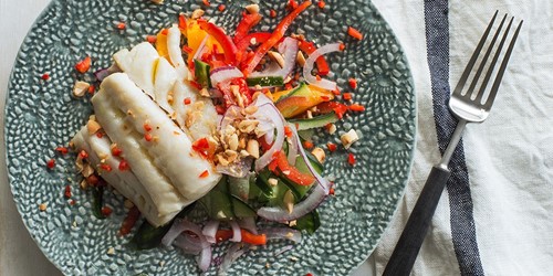 Steamed white fish fillets served over a vibrant Thai salad with sliced red onion, cucumber, capsicum, and crushed peanuts on a textured ceramic plate.