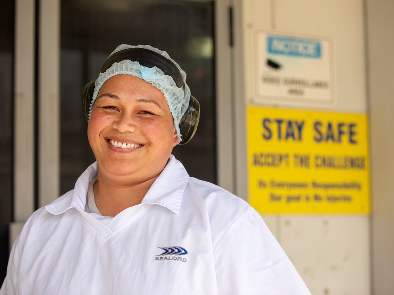 sealord factory worker standing in front of factory door smiling toward camera wearing sealord branded safety gear, hair net and air muffs