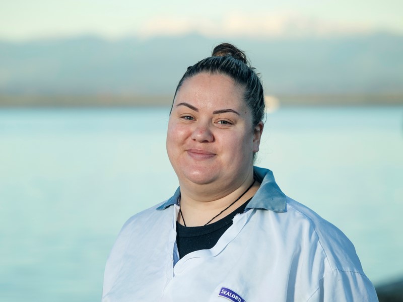 sealord worker standing infront of waterfront wearing a white sealord shirt and a black undershirt. has their hair tied back into a bun.