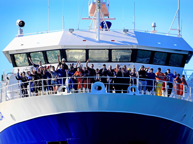 "Group of crew members standing and waving on the bow of a large commercial vessel under a clear blue sky.