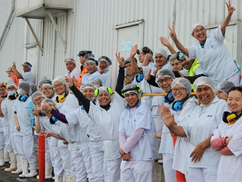 Large group of smiling factory workers wearing white uniforms, hairnets, and ear protection, gathered outside a building and waving at the camera.