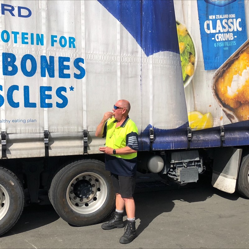 Man in a high-visibility vest eating a snack while standing beside a Sealord truck promoting New Zealand Hoki Classic Crumb fish fillets