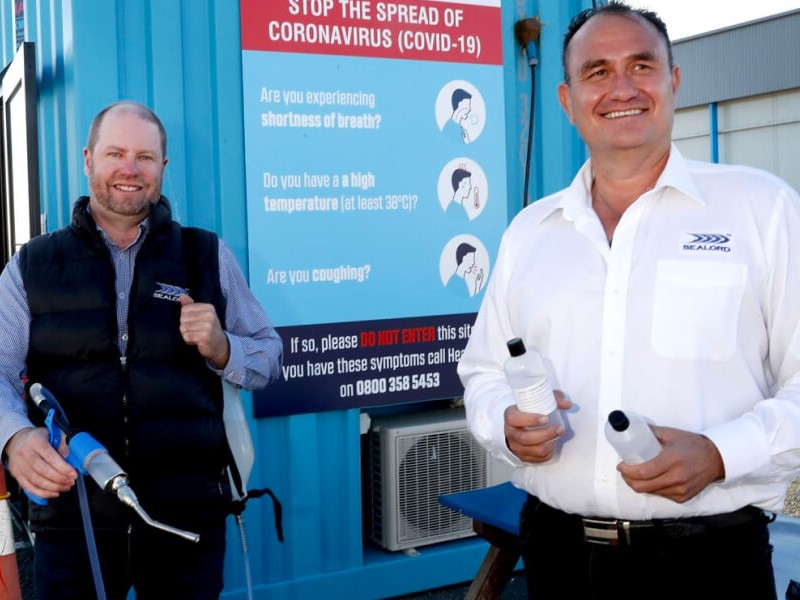 Sealord CEO and factory worker standing next to a COVID-19 safety sign; the worker holds a drill, and the CEO holds two water bottles, showcasing safe operations during Alert Level 3.
