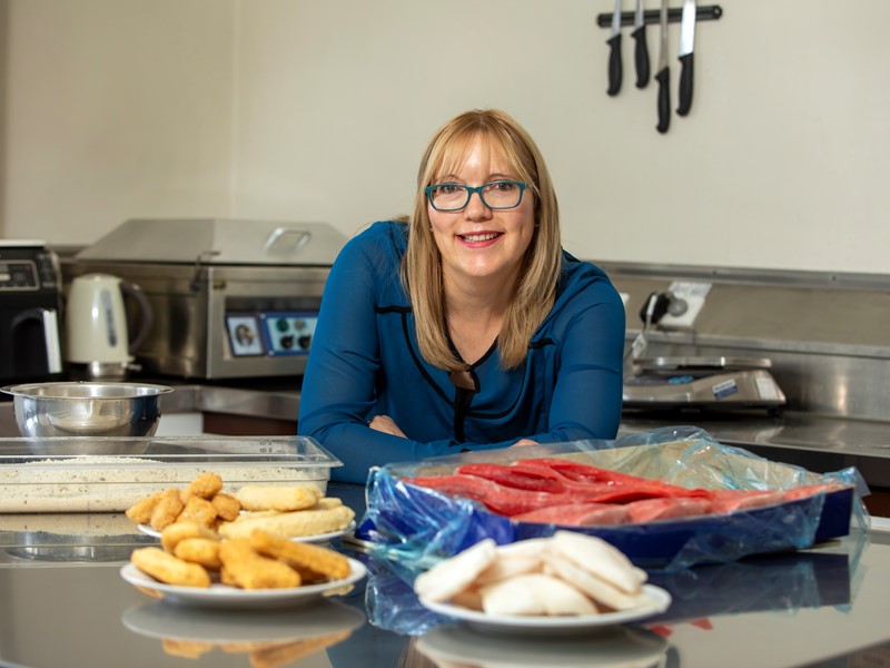 blonde women wearing glasses and a blue top leaning on a kitchen countertop with sealord products plated in front of her such as frozen crumbed fillets, bites, patties and fresh fish