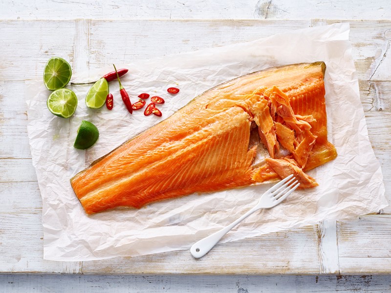Flaked smoked salmon fillet on parchment paper with lime halves and sliced red chili peppers, next to a white fork on a rustic wooden table.