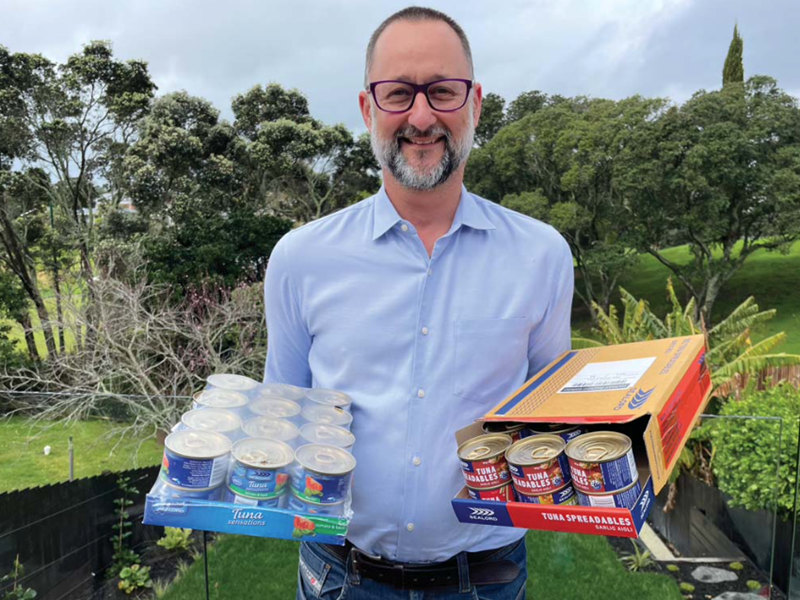 man wearing glasses, blue button up shirt and, salt and pepper beard holding 2 trays of canned tuna in front of a bright green garden