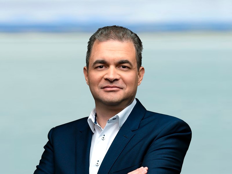 man with arms crossed in a white shirt and black blazer, mid shoy in front of the blue sea and sky looking toward the camera.
