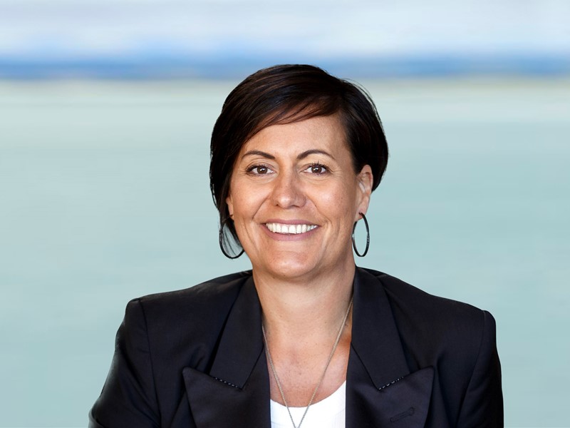 woman with pixie haircut , whoop earrings, black blazer and white shirt standing in front of the sea smiling towards camera
