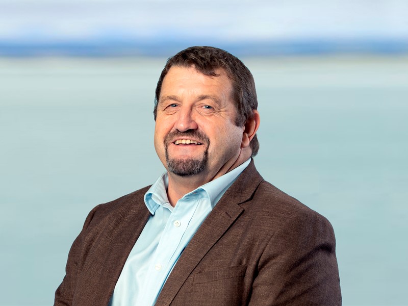 man standing in front of the sea and Mountains, with a brown blazer and blue shirt