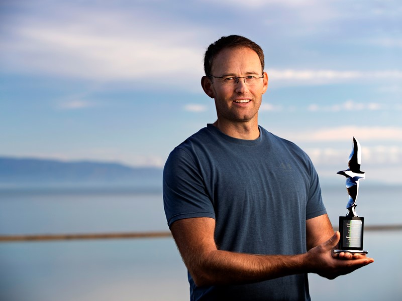 Man in a blue shirt holding a bird-shaped trophy, standing outdoors with the sea and mountains in the background under a blue sky.