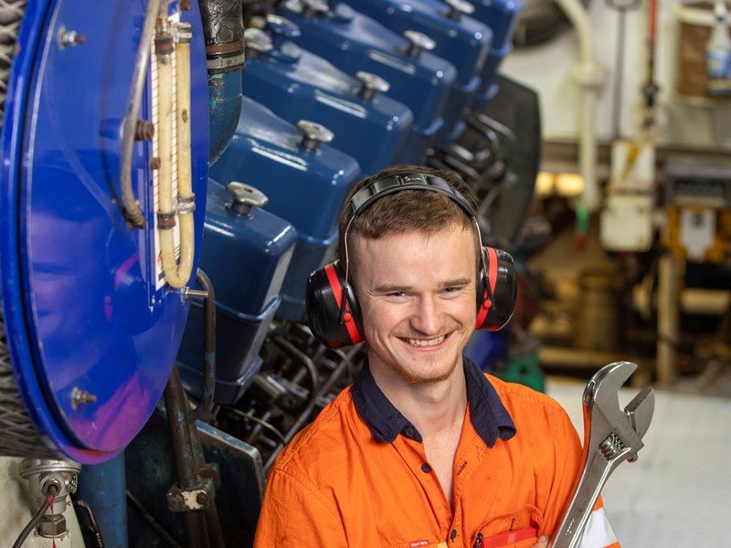 Smiling engineer wearing ear protection and an orange work shirt, holding a large wrench in front of industrial machinery