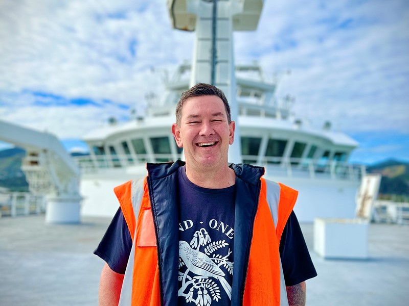man standing on front deck of sealords vessel laughing at the camera, wearing na blue and white t-shirt with a high visibility vest on, there is a blue sky with a few clouds above the man