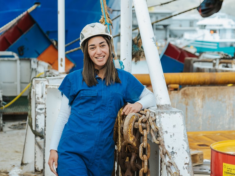 woman standing at the sealord doc near the vessel wearing blue overalls and a white safety hat leaning against a metal surface