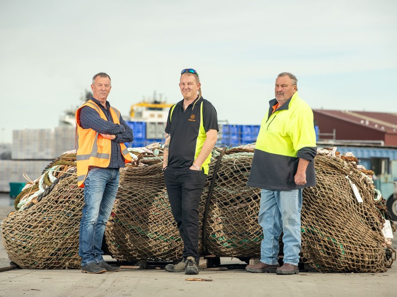 three Sealord workers standing in front of a large pile of old fishing nets on the dock with buildings and boats in the background promoting the company’s new recyclable fishing nets initiative