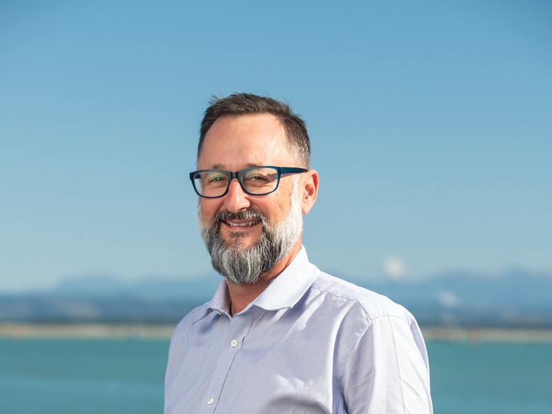 Smiling man with short dark hair, glasses, and a salt-and-pepper beard, wearing a light purple collared shirt, standing in front of a clear blue sky and ocean background