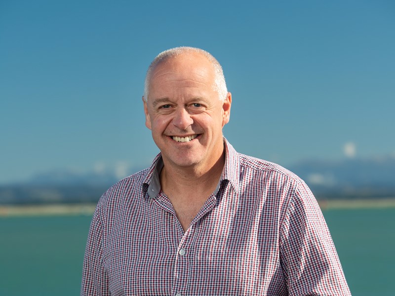 Smiling man with short grey hair wearing a red and white checkered shirt, standing in front of a clear blue sky and ocean backdrop.