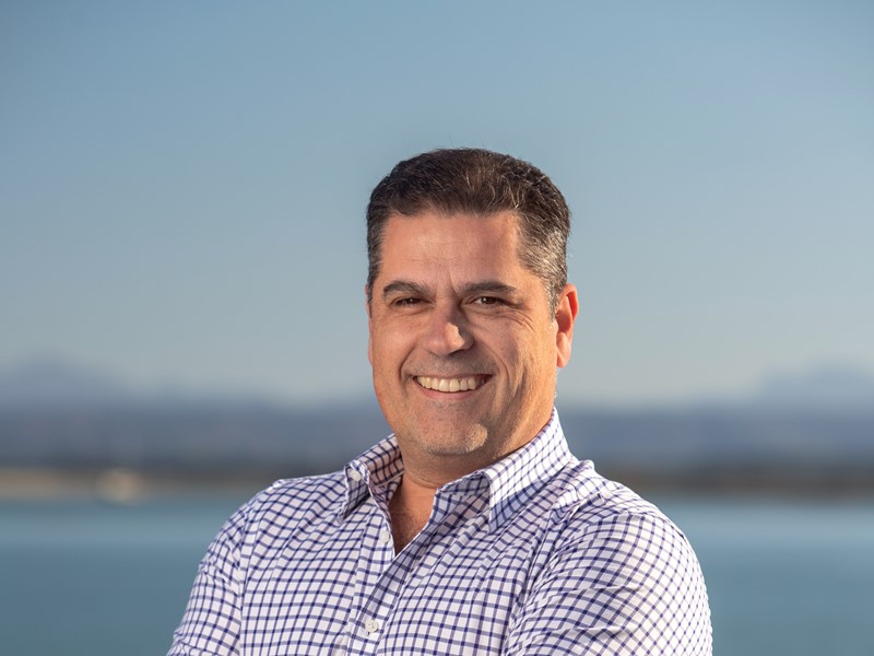 Smiling man with short dark hair wearing a white shirt with blue checks, standing with arms crossed in front of a clear sky and ocean backdrop