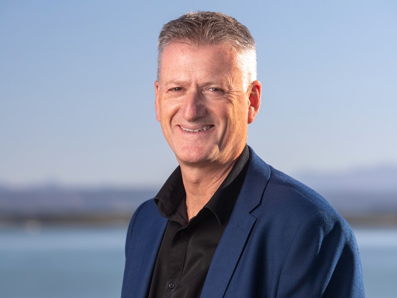 Smiling man with short grey hair wearing a dark shirt and blue blazer, standing in front of a clear sky and water backdrop