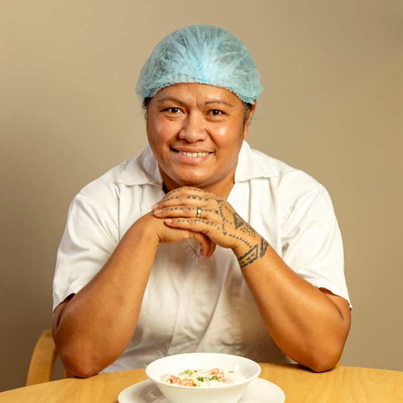 Smiling food worker wearing a blue hair net and white uniform, sitting at a table with a bowl of coconut fish salad.