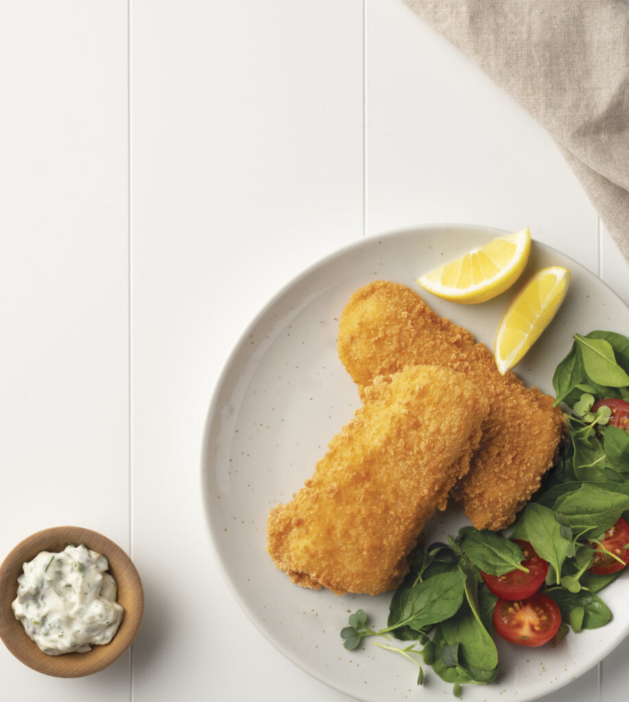 Plate with two golden crumbed fish fillets, fresh spinach and cherry tomato salad, and lemon wedges. A small wooden bowl of tartar sauce sits beside the plate on a white table with a beige napkin.