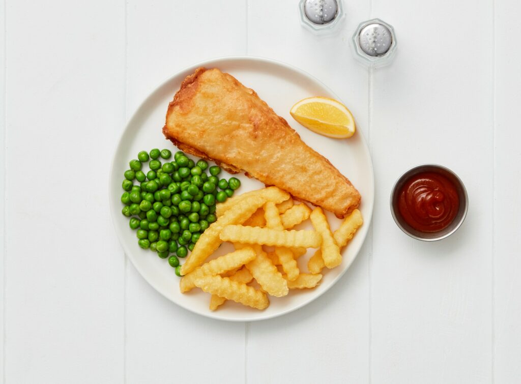 Classic fish and chips meal with battered Hoki portion, crinkle-cut fries, green peas, a lemon wedge, and ketchup, served on a white plate.